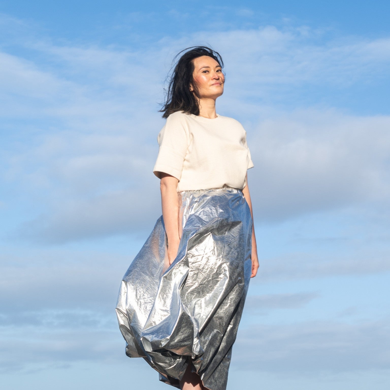 Woman wearing a white top and silver metallic skirt standing against a blue sky with clouds.
