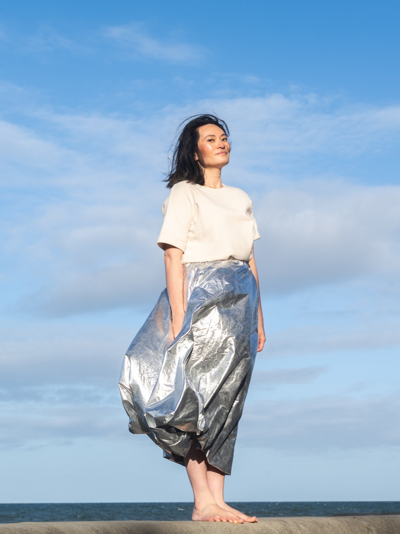 Woman wearing a white top and silver metallic skirt standing against a blue sky with clouds.